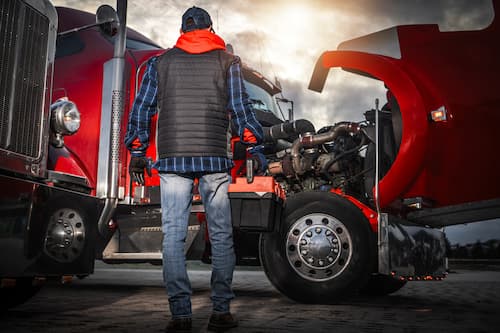 Man in a vest and hoodie inspecting the engine of two red semi-trucks with their hoods raised.
